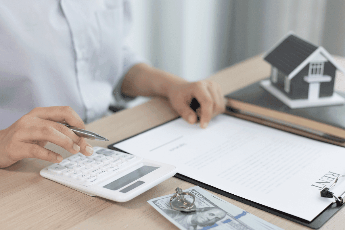 A woman signs a contract, calculating figures with a house model nearby, ensuring a secure transaction.