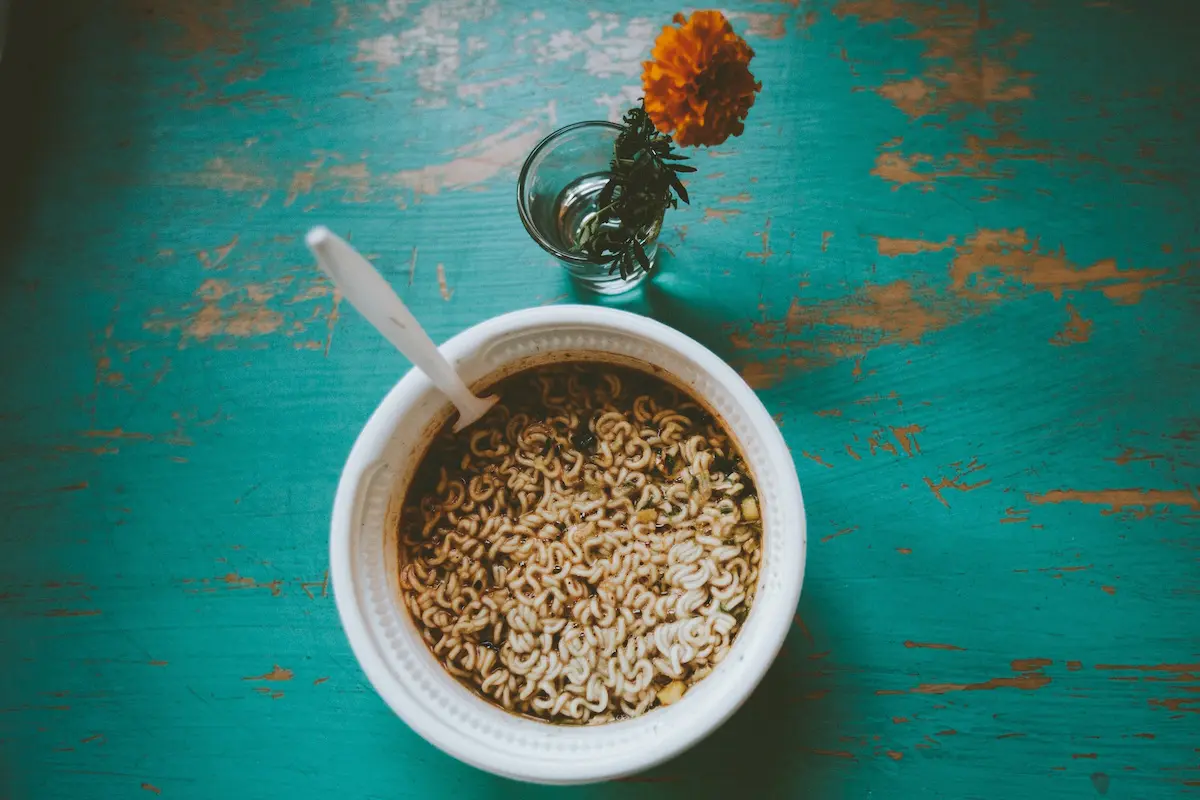 A wooden table hosts a bowl of noodles and a small glass of water next to a flower, creating a wholesome breakfast scene.