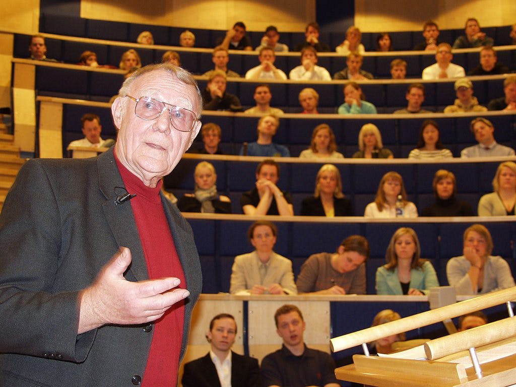 An older man stands before a lecture hall, surrounded by a group of people, engaged in a discussion or presentation.