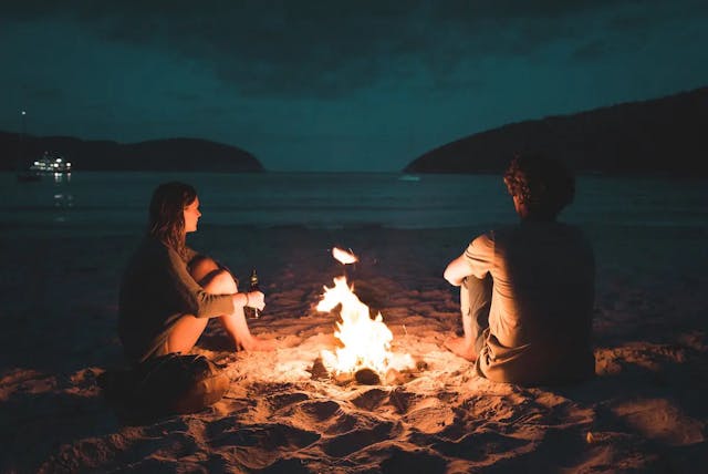 a couple sitting around a fire on a beach at night