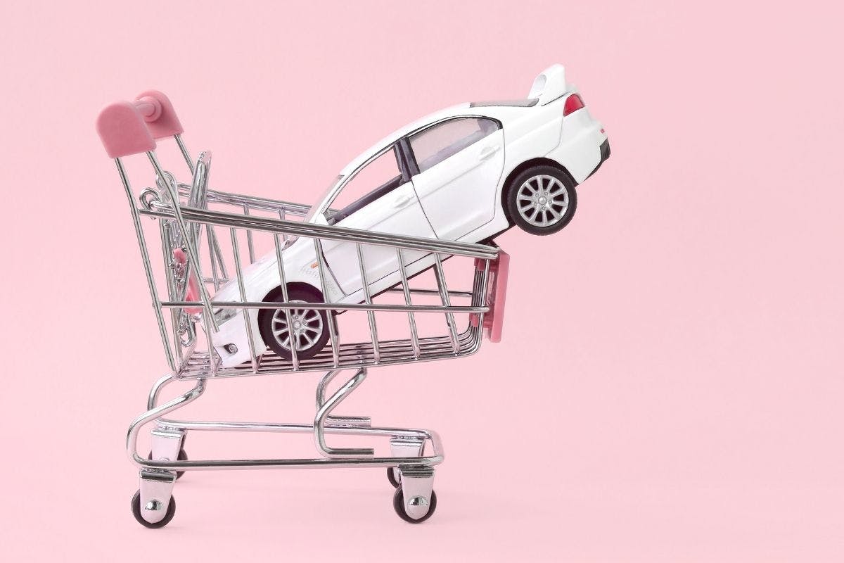 A car placed inside a shopping cart against a pink backdrop, showcasing a unique combination of transportation and shopping elements.