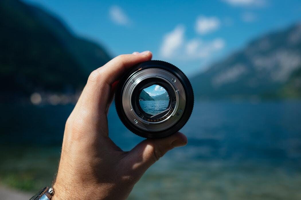 a hand holding a camera lens with a lake in the background