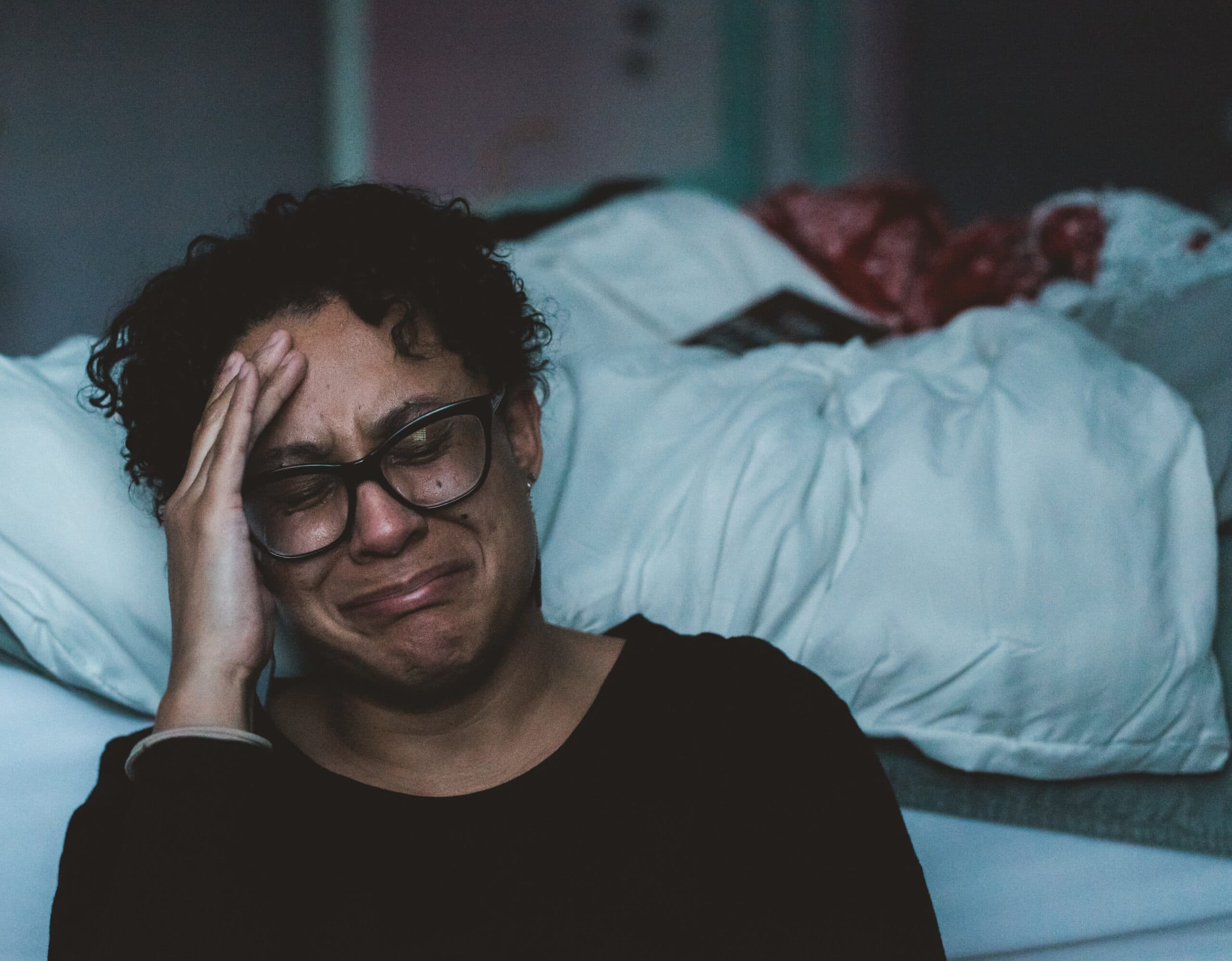 A woman wearing glasses sits on a bed, her head resting in her hands.