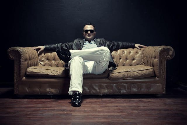 A man seated on a leather couch against a black wall, exuding comfort and style.