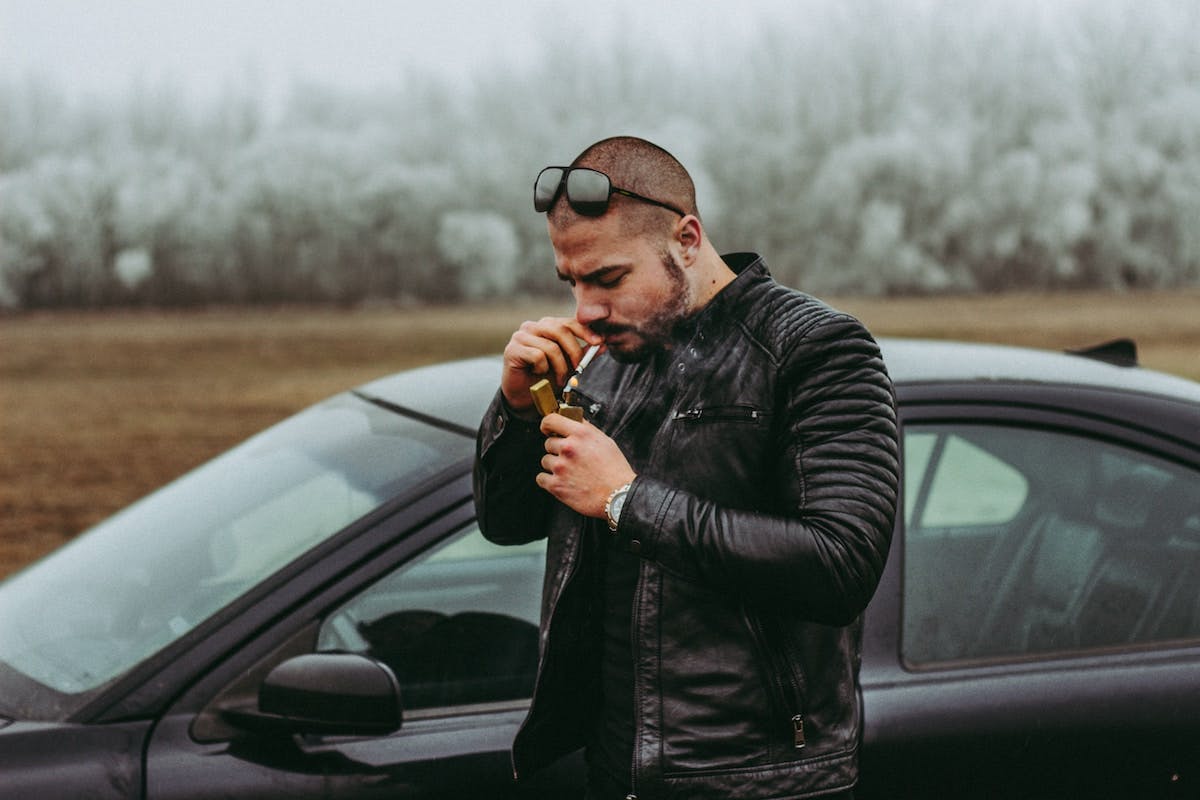 A man in a leather jacket smoking a cigarette by a car, exuding a cool and rebellious aura.